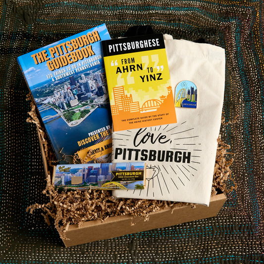 A curated gift box containing a blue 'The Pittsburgh Guidebook', a yellow and black 'Pittsburghese' book, and a white tote printed with 'love, PITTSBURGH' and a skyline graphic, arranged in brown paper shred on a dark woven blanket.