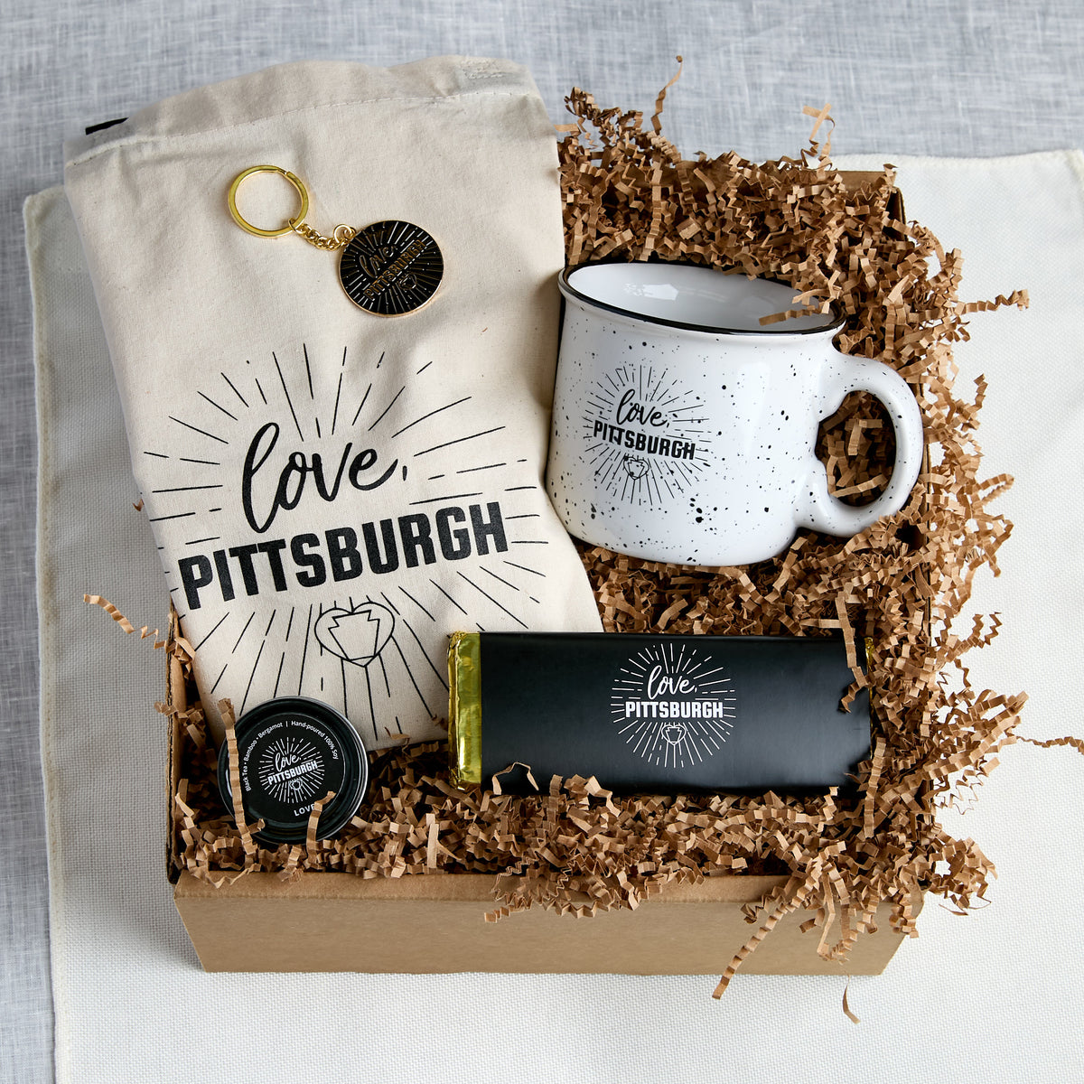 A gift box set with 'Love Pittsburgh' themed items, including a canvas tote bag, a speckled enamel mug, a chocolate bar, and a small canlde tin, arranged in a box with brown paper shred.