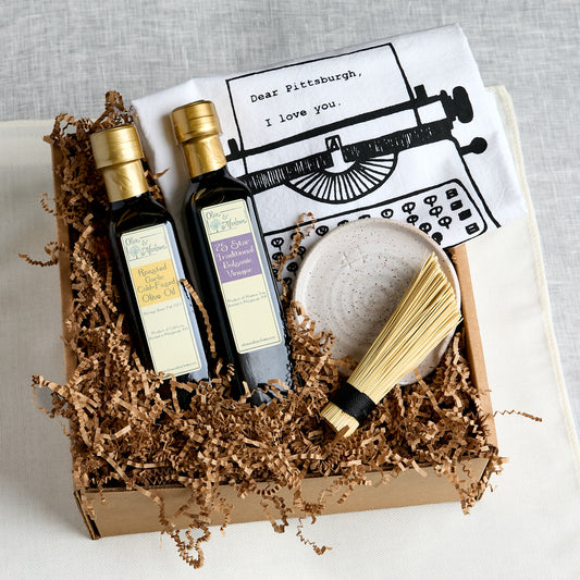 A curated gift box set containing two bottles of Olive and Marlow Olive Oil, and Balsamic vinegar, a white tea towel with a typewriter graphic and 'Dear Pittsburgh, I love you.' text, a ceramic spoon rest, and a pot scrubber, all arranged in brown paper shred inside a cardboard box.