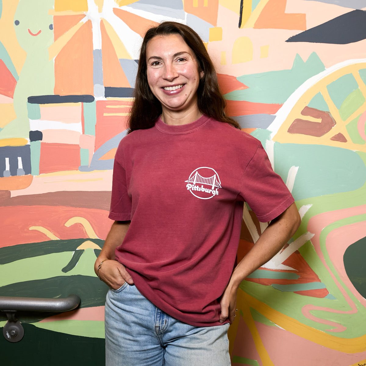 A woman smiling and posing in front of a colorful abstract mural, wearing a faded deep red t-shirt with a white 'Pittsburgh' logo on the chest and light wash blue jeans.