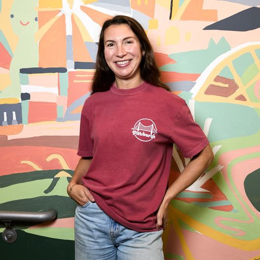 A woman smiling and posing in front of a colorful abstract mural, wearing a faded deep red t-shirt with a white 'Pittsburgh' logo on the chest and light wash blue jeans.