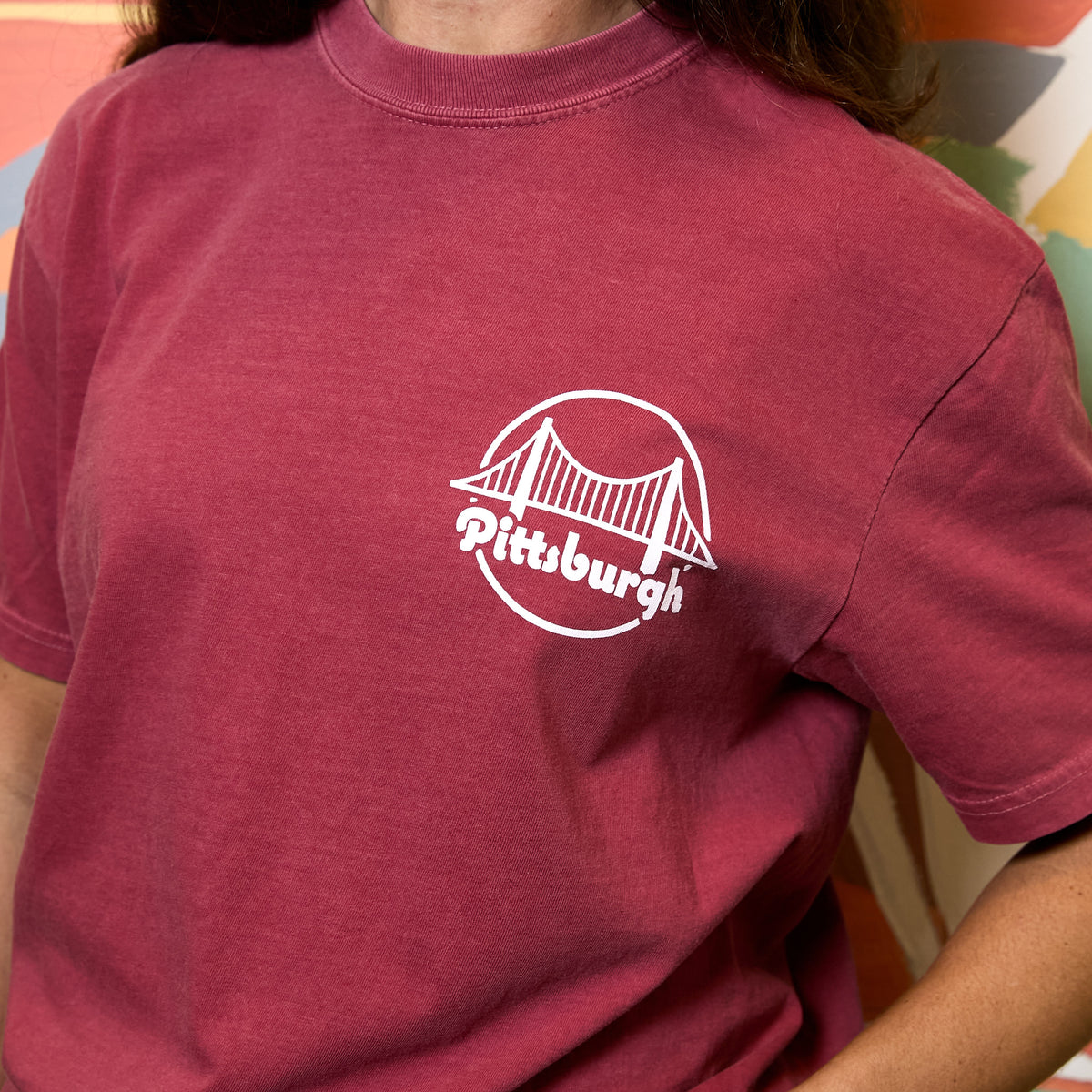 A person wearing a washed deep red t-shirt with a white circular Pittsburgh bridge graphic on the left chest.