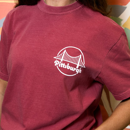 A person wearing a washed deep red t-shirt with a white circular Pittsburgh bridge graphic on the left chest.