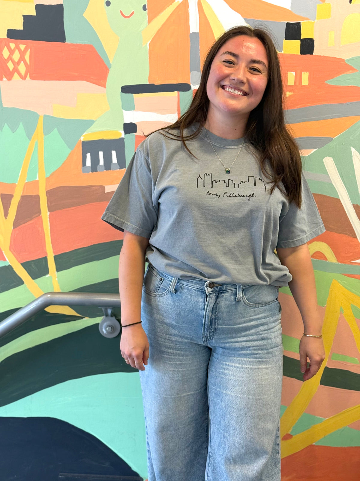A smiling young woman stands indoors in front of a colorful mural, wearing a light gray crewneck t-shirt with a black Pittsburgh skyline graphic and text, paired with light-wash blue jeans.