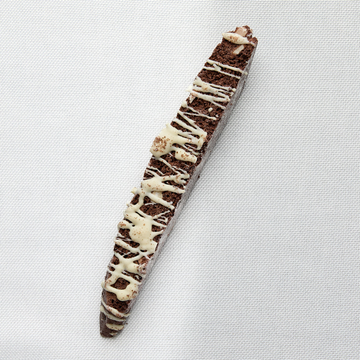 A chocolate biscotti with white chocolate drizzle, photographed diagonally on a white textured fabric background.