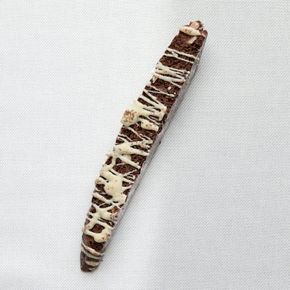 A chocolate biscotti with white chocolate drizzle, photographed diagonally on a white textured fabric background.