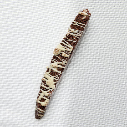 A chocolate biscotti with white chocolate drizzle, photographed diagonally on a white textured fabric background.