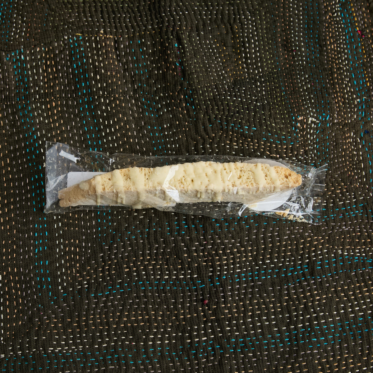 A Biscotti - White Chocolate Macadamia Nut, wrapped in clear plastic packaging and placed on a dark woven mat with blue and beige stitching.