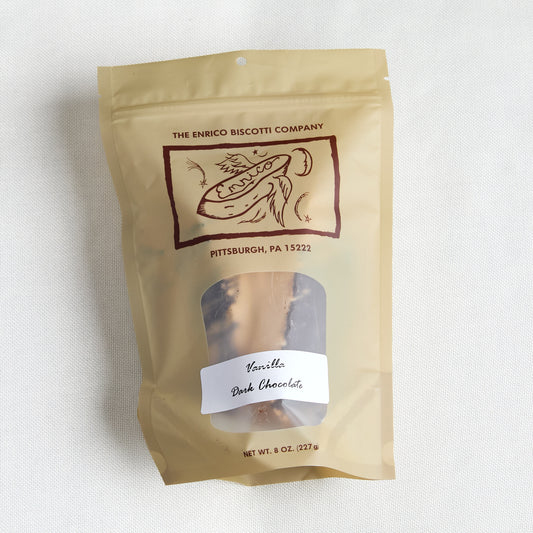 A stand-up pouch of The Enrico Biscotti Company Vanilla Dark Chocolate on a light textured fabric background. The pouch is a warm tan color with a clear window showing the chocolate inside and a white label on the front.