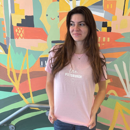Person wearing a pink 'love, Pittsburgh' logo t-shirt standing in front of a colorful mural.