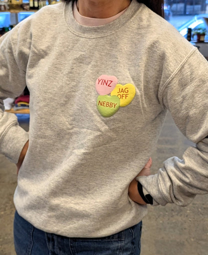 Person wearing a light gray sweatshirt with colorful heart-shaped puffy print Pittsburghese conversation hearts that say "nebby" "yinz" and "jagoff"