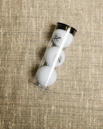 Three golf balls in a clear cylindrical container on a textured beige surface