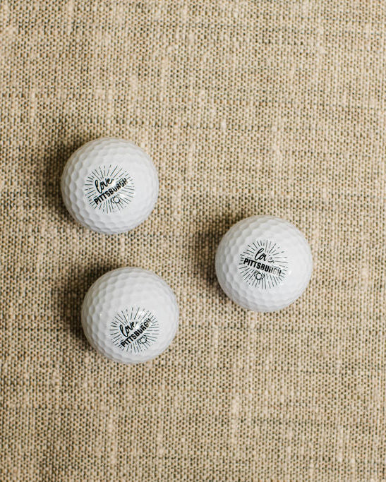 Three white golf balls with a logo on a beige fabric background