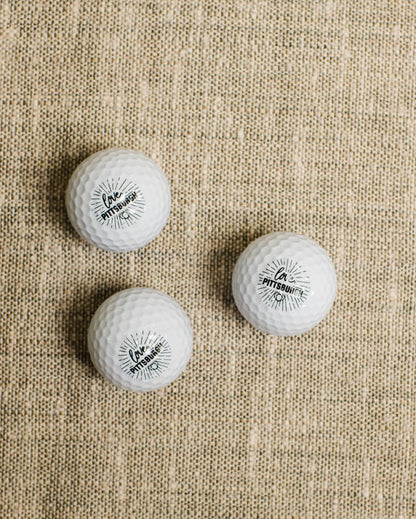 Three white golf balls with a logo on a beige fabric background