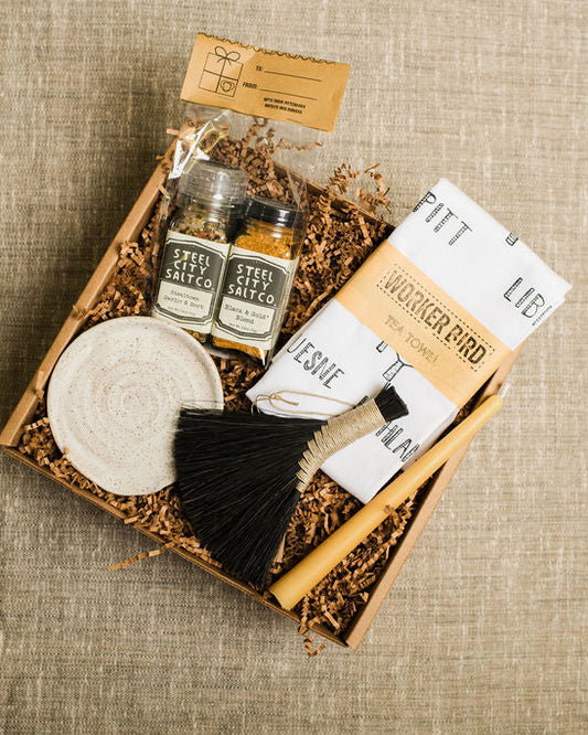 Gift set for the home with salts, tea towel, taper candle, small broom, and spoon rest on a textured surface
