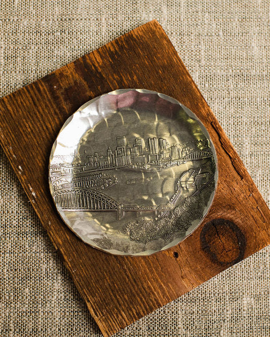 aluminum coaster with pittsburgh skyline on a wooden block with a textured beige background