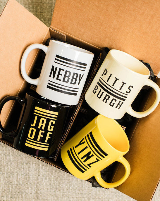 Four pittsburgh mugs with text designs in a box on a textured surface
