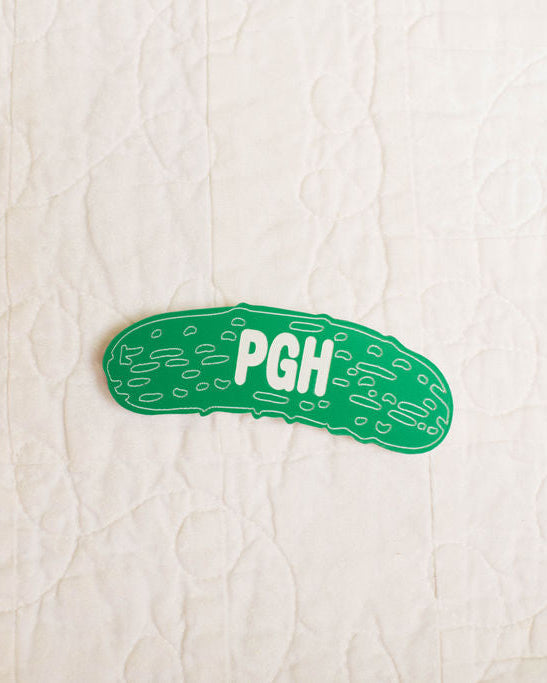Green pickle-shaped magnet with 'PGH' text on a textured beige background