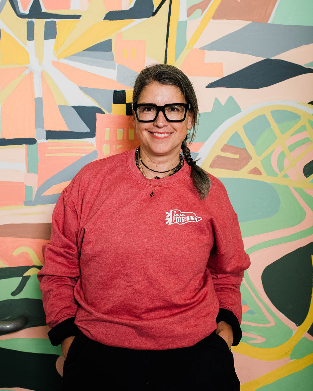 Person wearing a red sweatshirt with a logo in front of a colorful abstract mural.