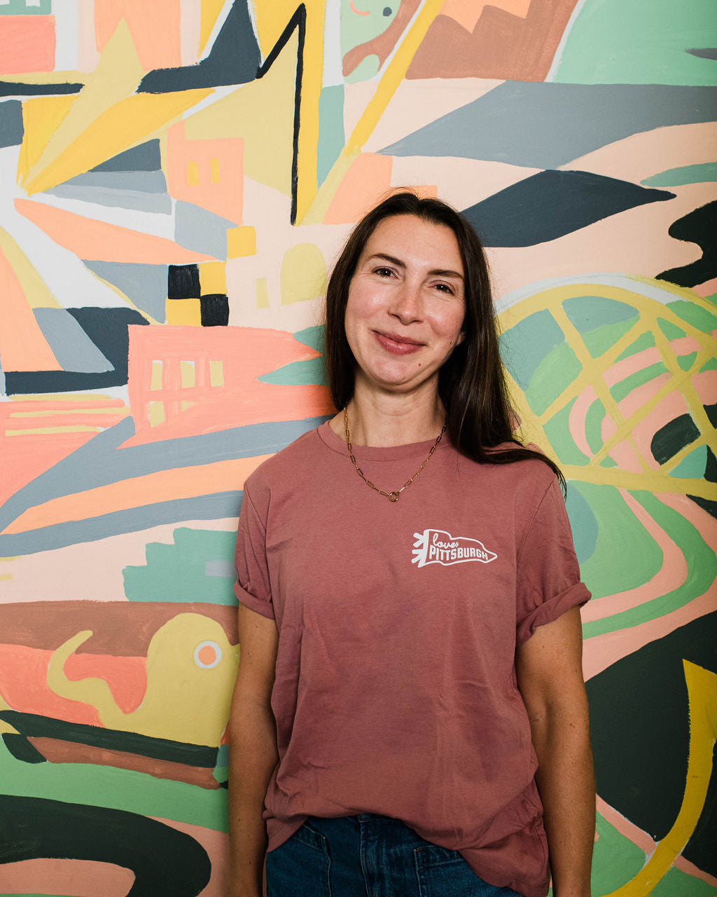Woman wearing a maroon t-shirt with a logo in front of a colorful abstract wall mural.
