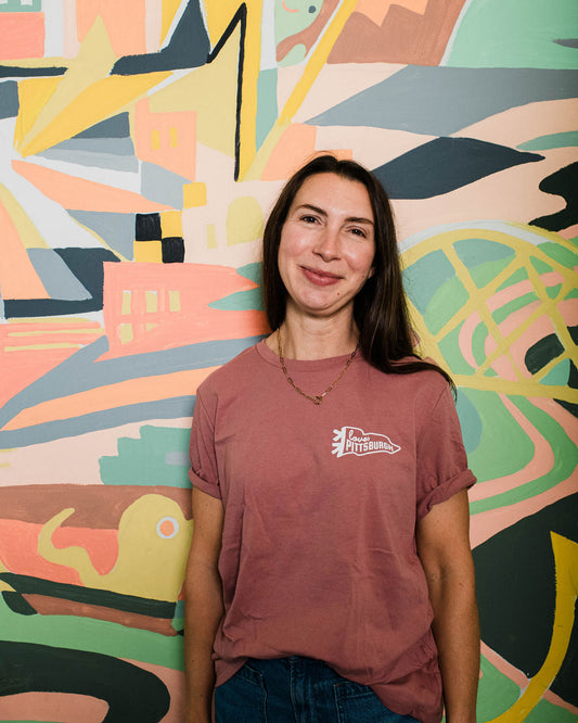 Woman wearing a maroon t-shirt with a logo in front of a colorful abstract wall mural.