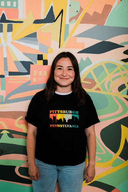 Person wearing a black t-shirt with 'Pittsburgh Pennsylvania' design in front of a colorful abstract wall.
