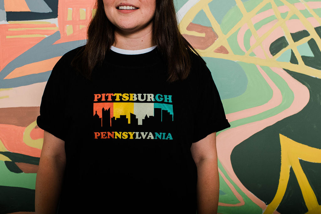 Person wearing a black t-shirt with 'Pittsburgh Pennsylvania' design in front of a colorful mural.
