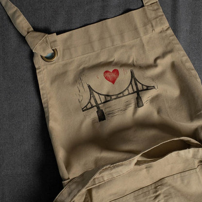 A beige apron featuring a black and red heart over a bridge block print design with a red heart in the center.